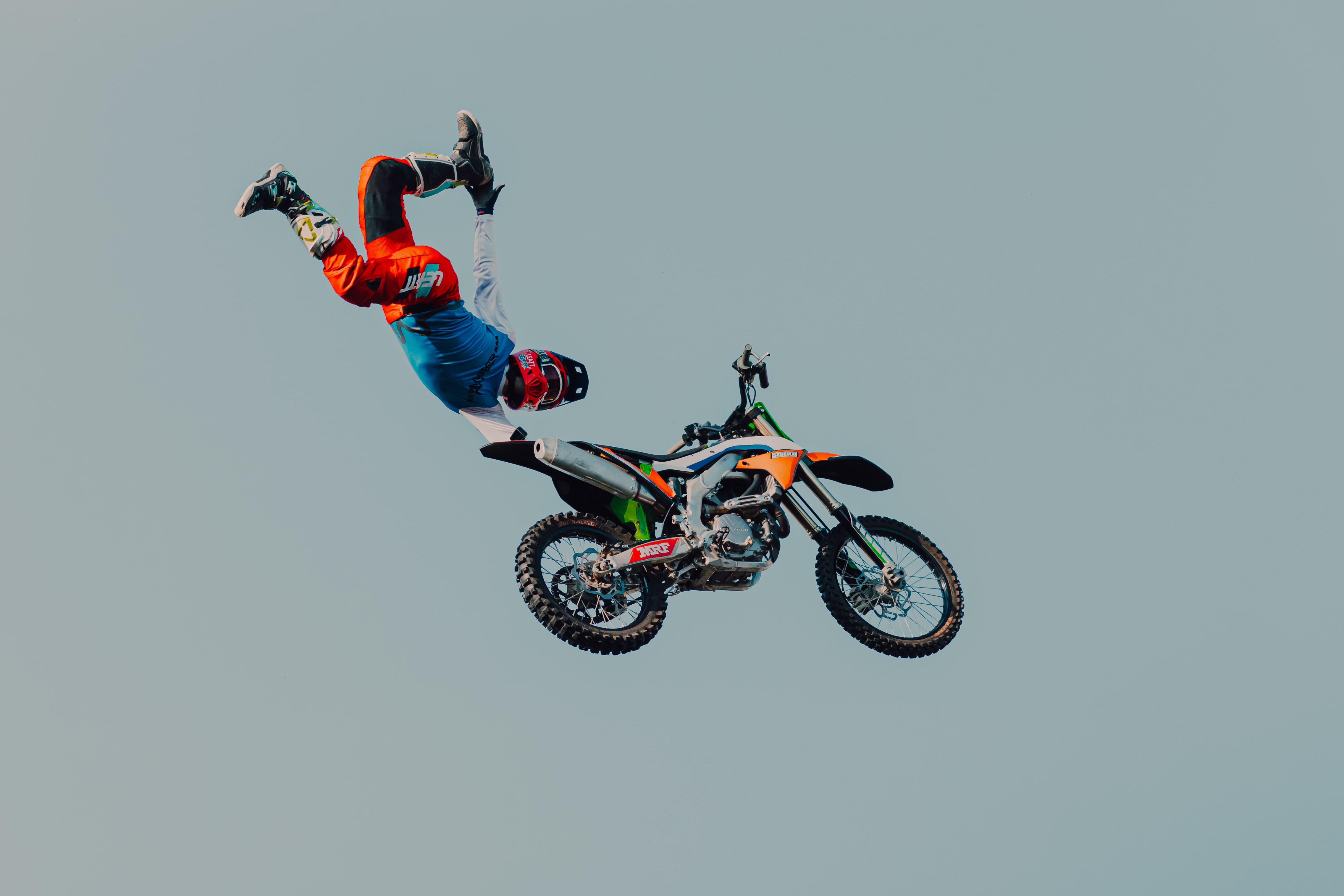 Motorcyclist in orange and blue gear performing a mid-air stunt away from the motorcycle against a clear sky.
