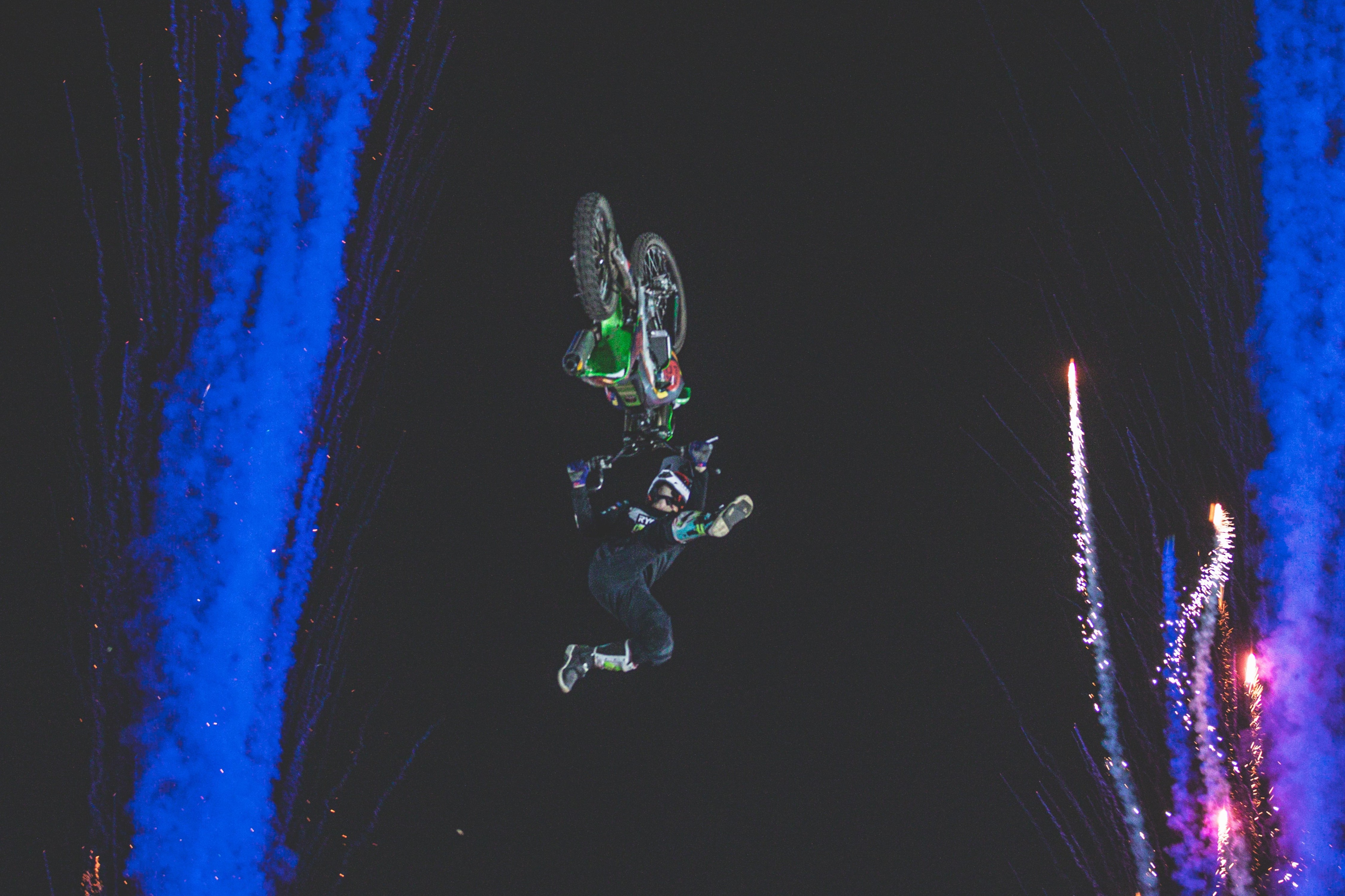 Motorcyclist performing a backflip stunt on a dirt bike at night with blue smoke and fireworks in the background.