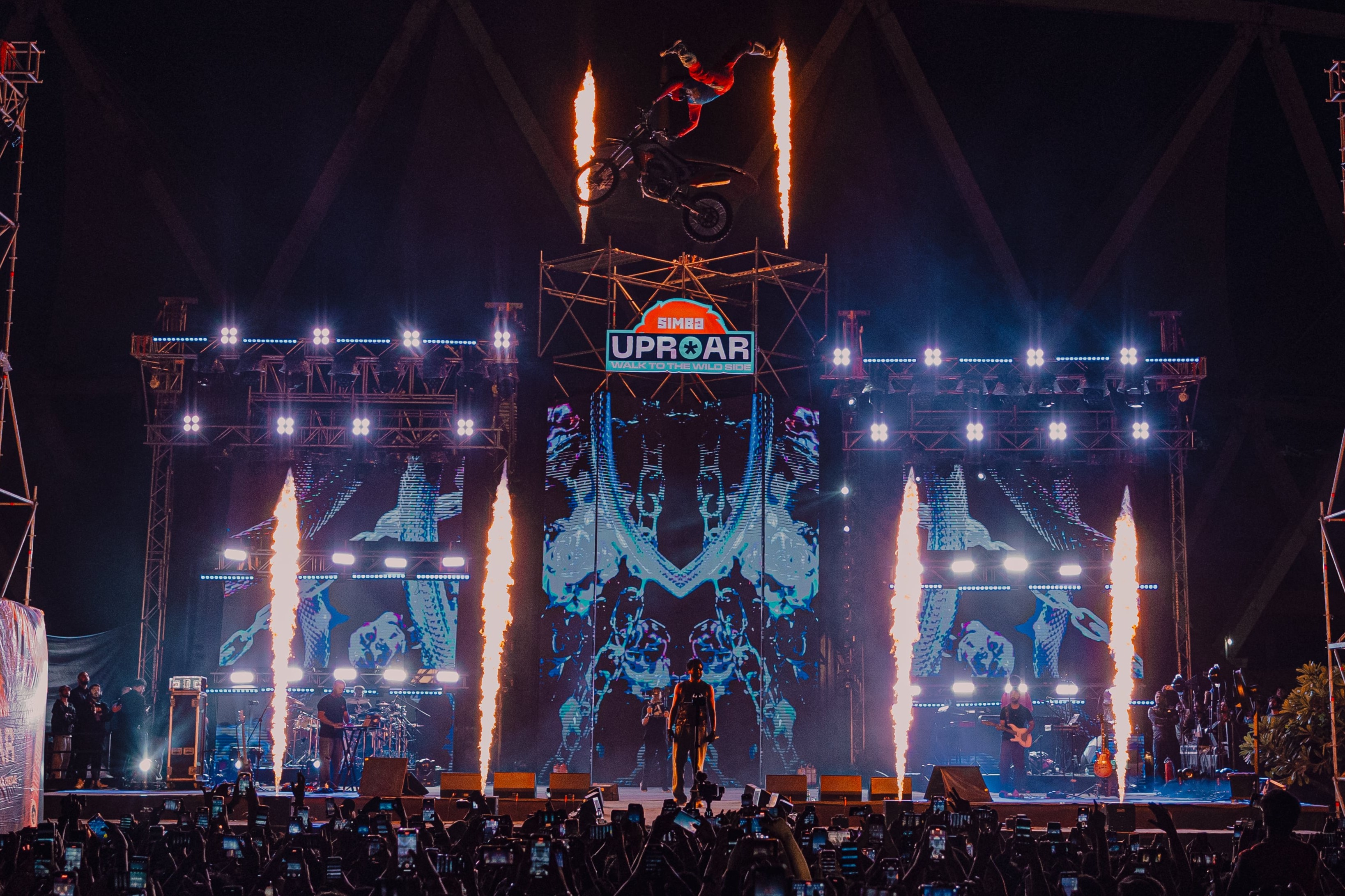 Motorcyclist performing a mid-air stunt above a stage with flames and bright lights during Simba Uproar event.