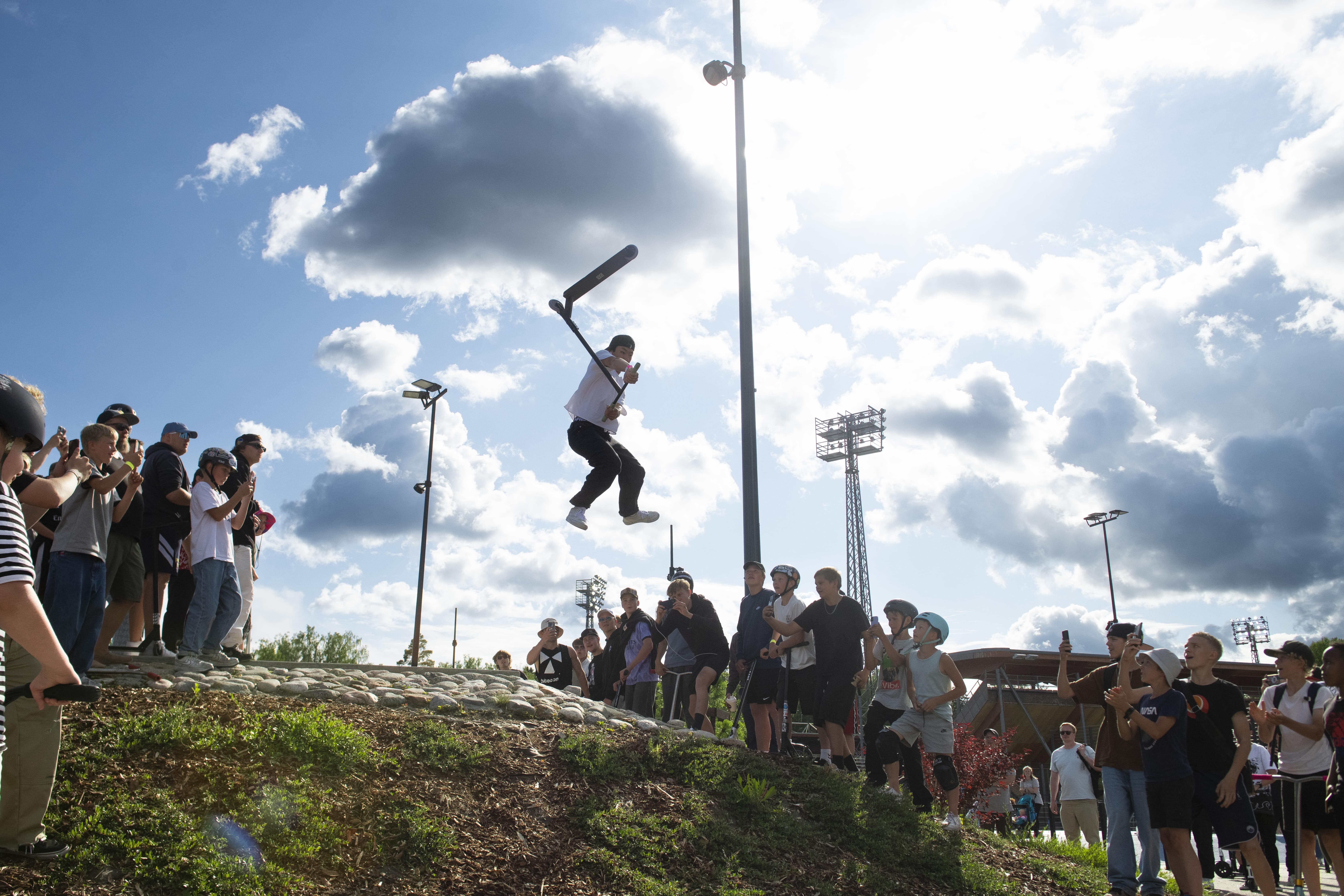 A person mid-air while performing a stunt on a scooter as a crowd of spectators watches and records under a partly cloudy sky.