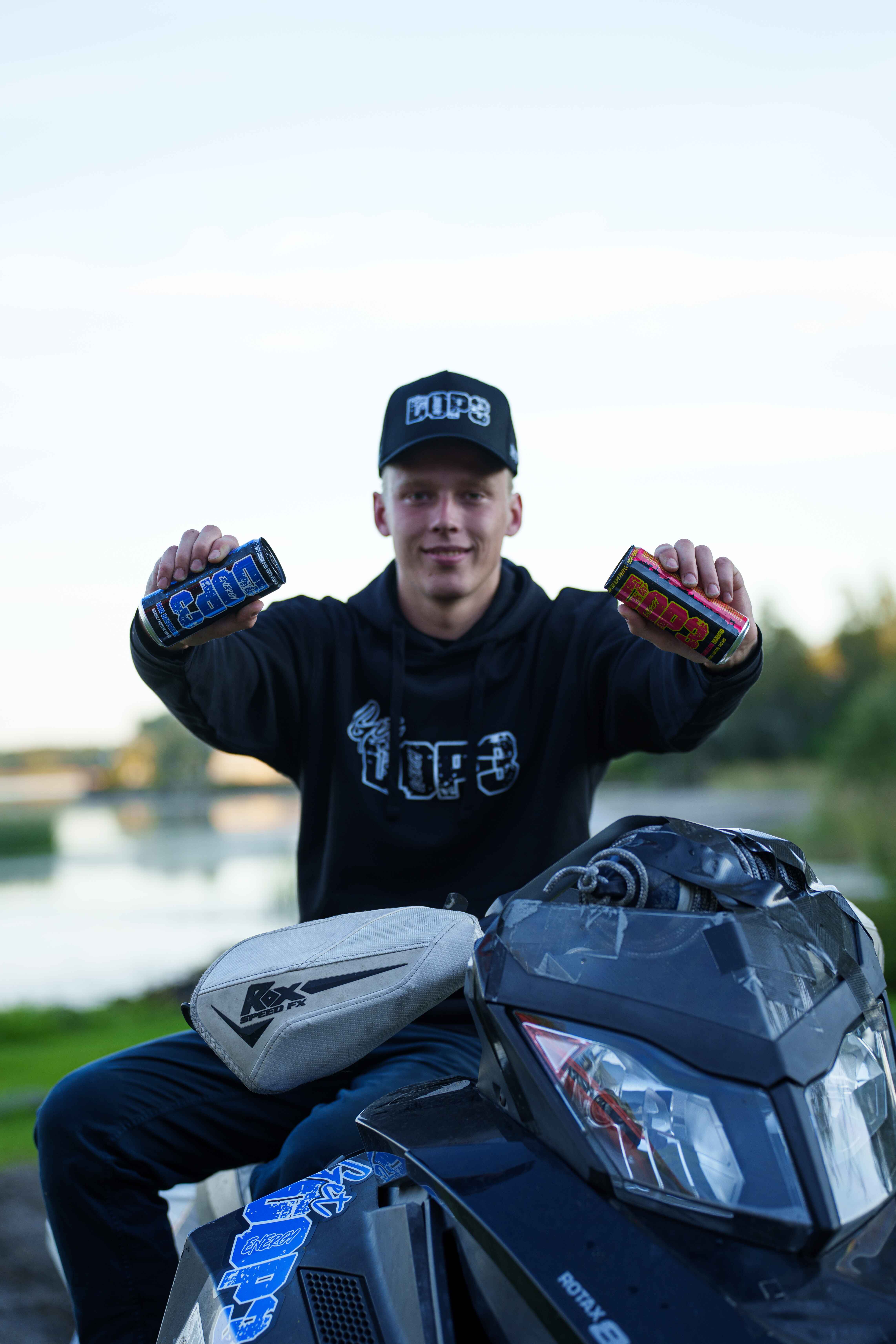 Young man wearing a black cap and hoodie holding blue and red energy drink cans, sitting on a black snowmobile outdoors near a body of water.