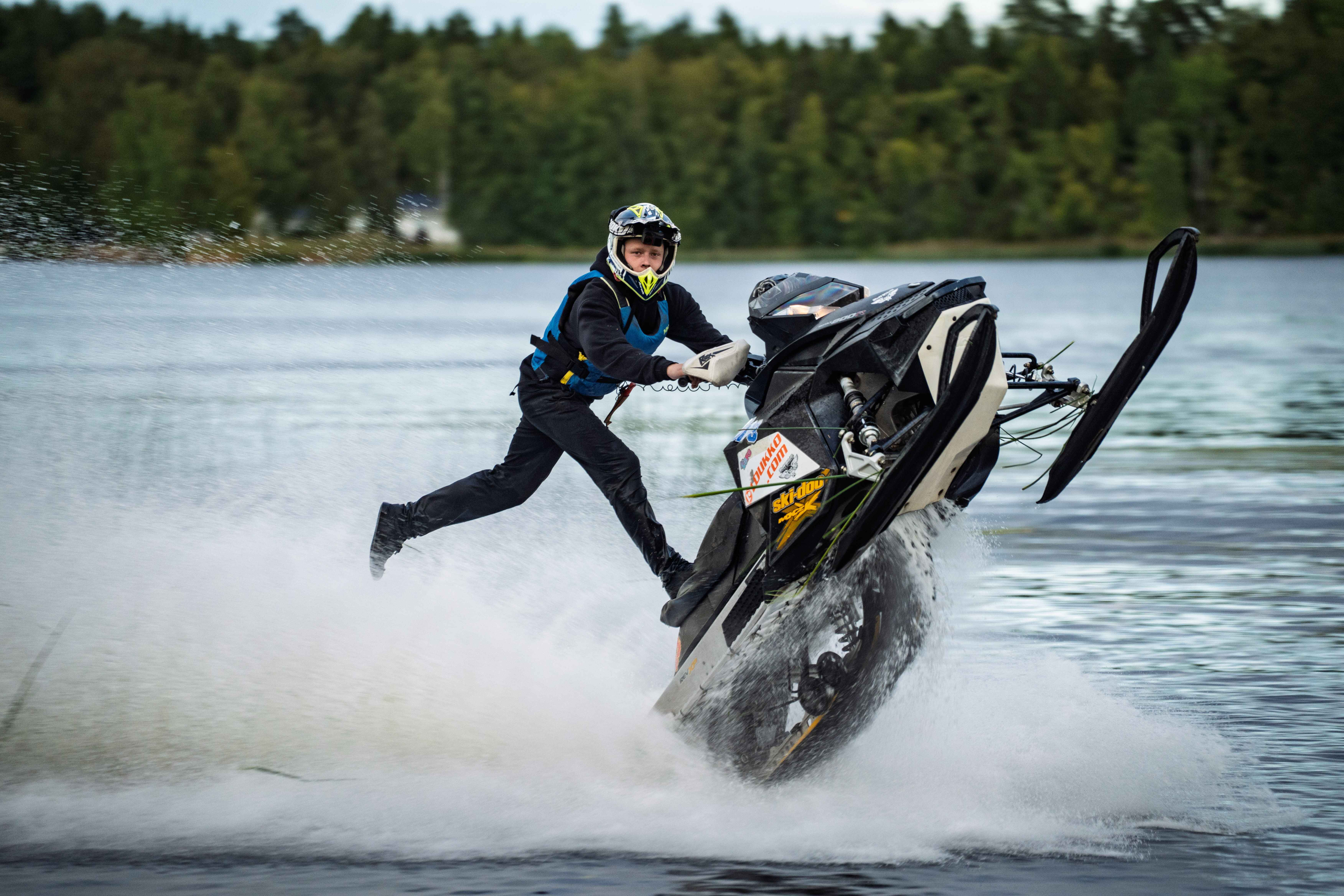 Person in helmet and life vest performing a stunt by standing on one leg while riding a snowmobile on water with a splash effect.