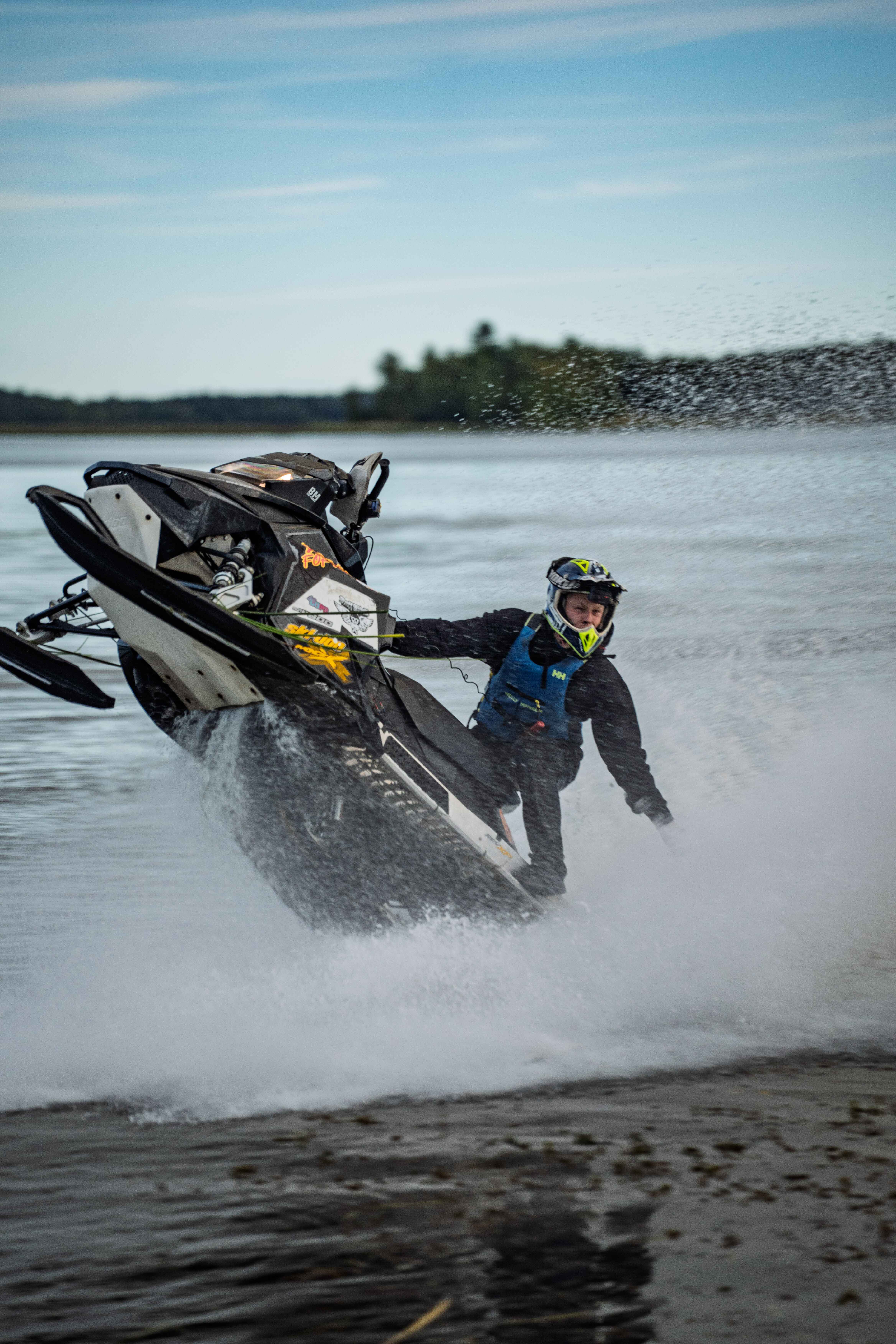 Person in protective gear performing a high jump stunt on a black snowmobile over water, splashing water around.