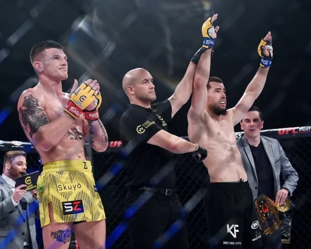 MMA referee raising the arm of the victorious fighter inside the octagon while the opponent claps.