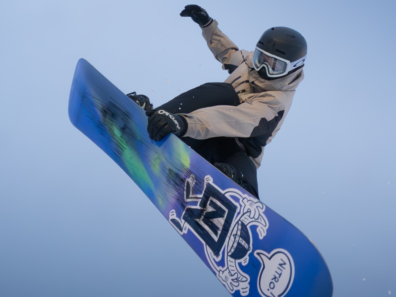 Snowboarder wearing beige jacket, black helmet, and goggles performing an aerial trick with a blue snowboard featuring a cartoon graphic and the word 'NITRO.'