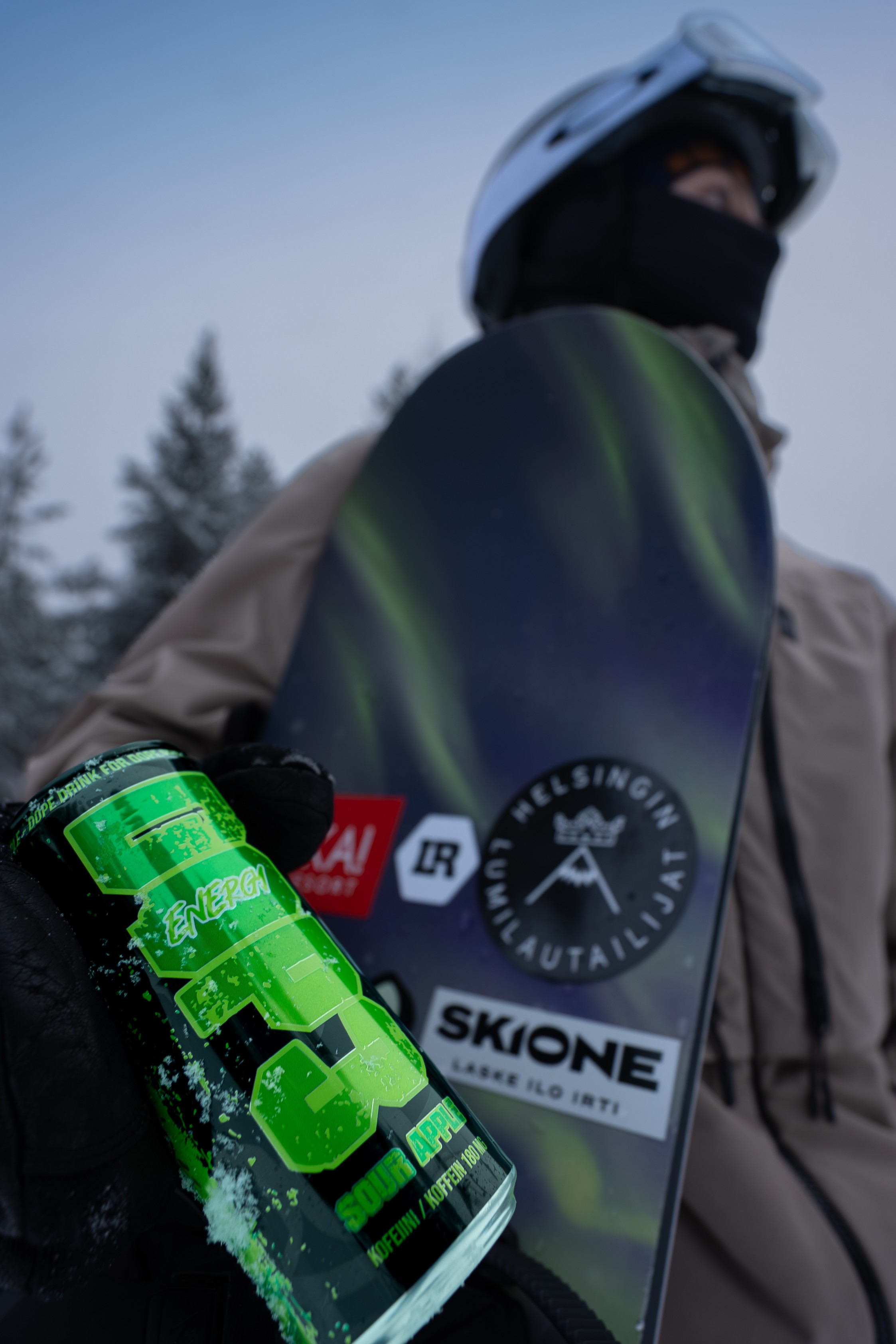 Person holding a bright green sour apple energy drink can and a snowboard with stickers, wearing winter gear and a helmet.