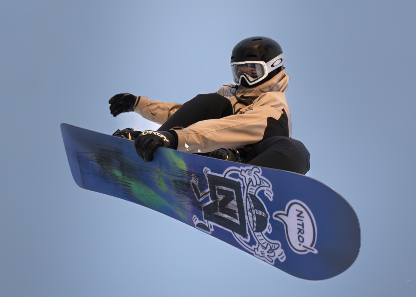 Snowboarder in beige jacket and black helmet performing an aerial trick against a clear blue sky.