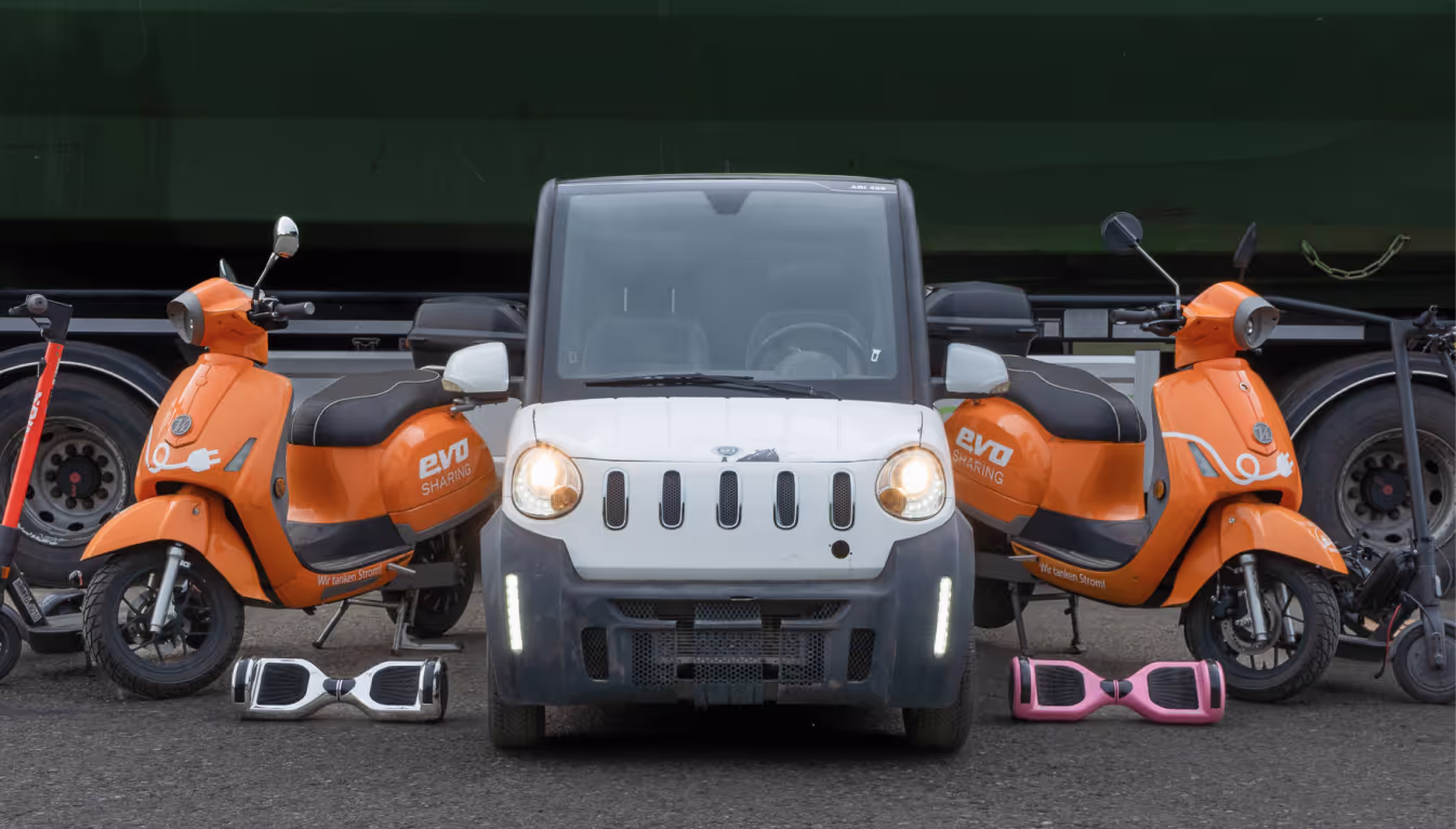 Verschiedene elektronische Fortbewegungsmittel, E-Roller, Scooter und ein Auto stehen vor einem grünen Container