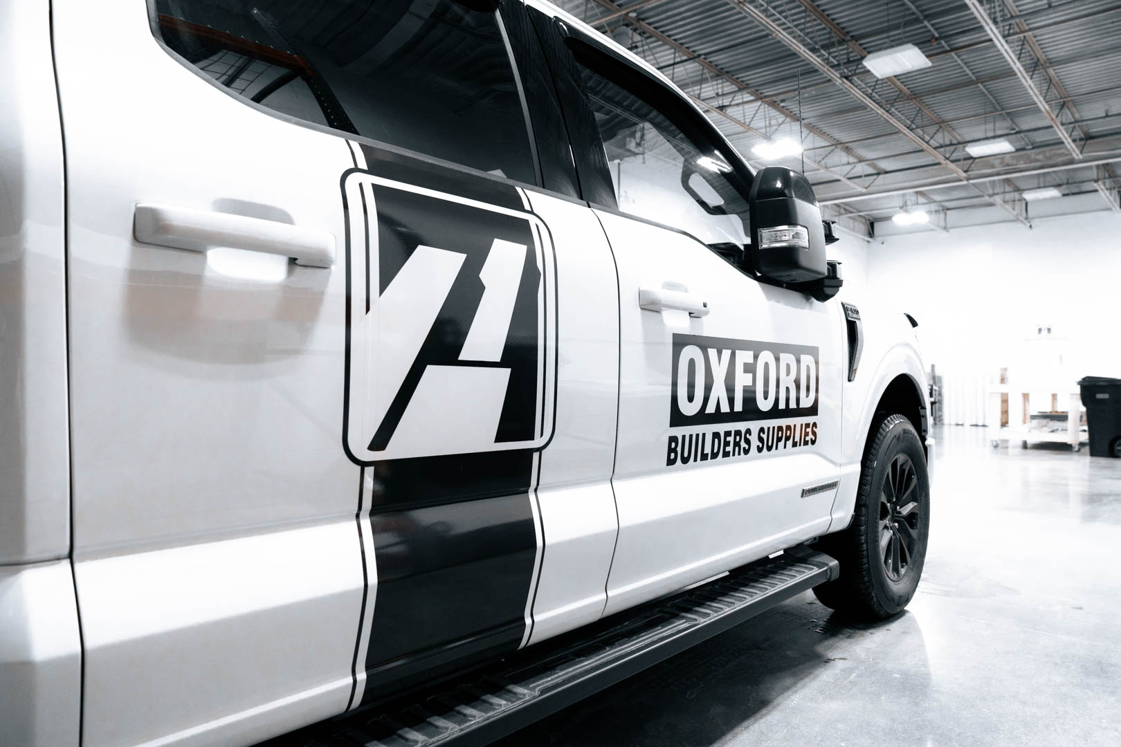 Side view of a white truck with black decal and text reading 'OXFORD BUILDERS SUPPLIES' parked inside a warehouse.