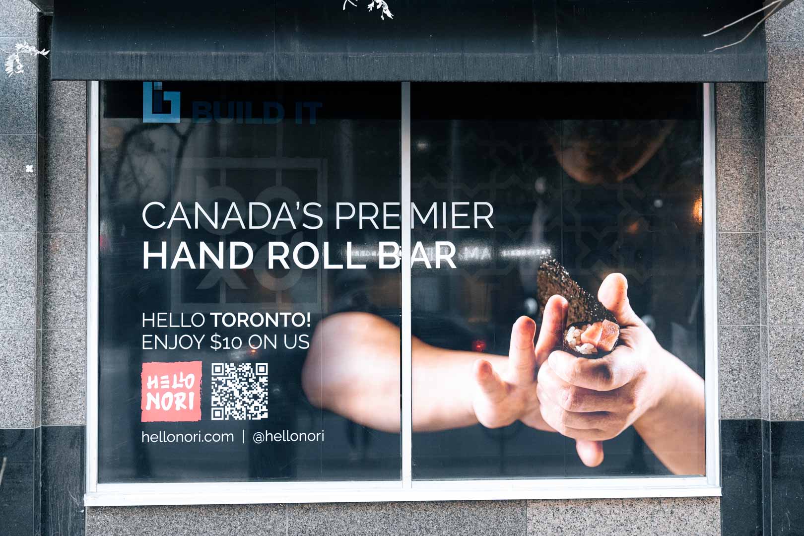 Window advertisement for Hello Nori, Canada's premier hand roll bar in Toronto, showing hands preparing a sushi hand roll.