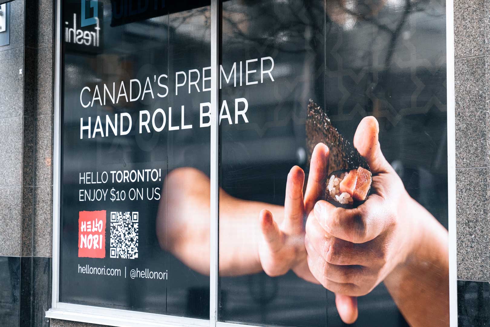 Window advertisement for Canada's premier hand roll bar showing hands holding a sushi hand roll and promotion offering $10 off in Toronto.