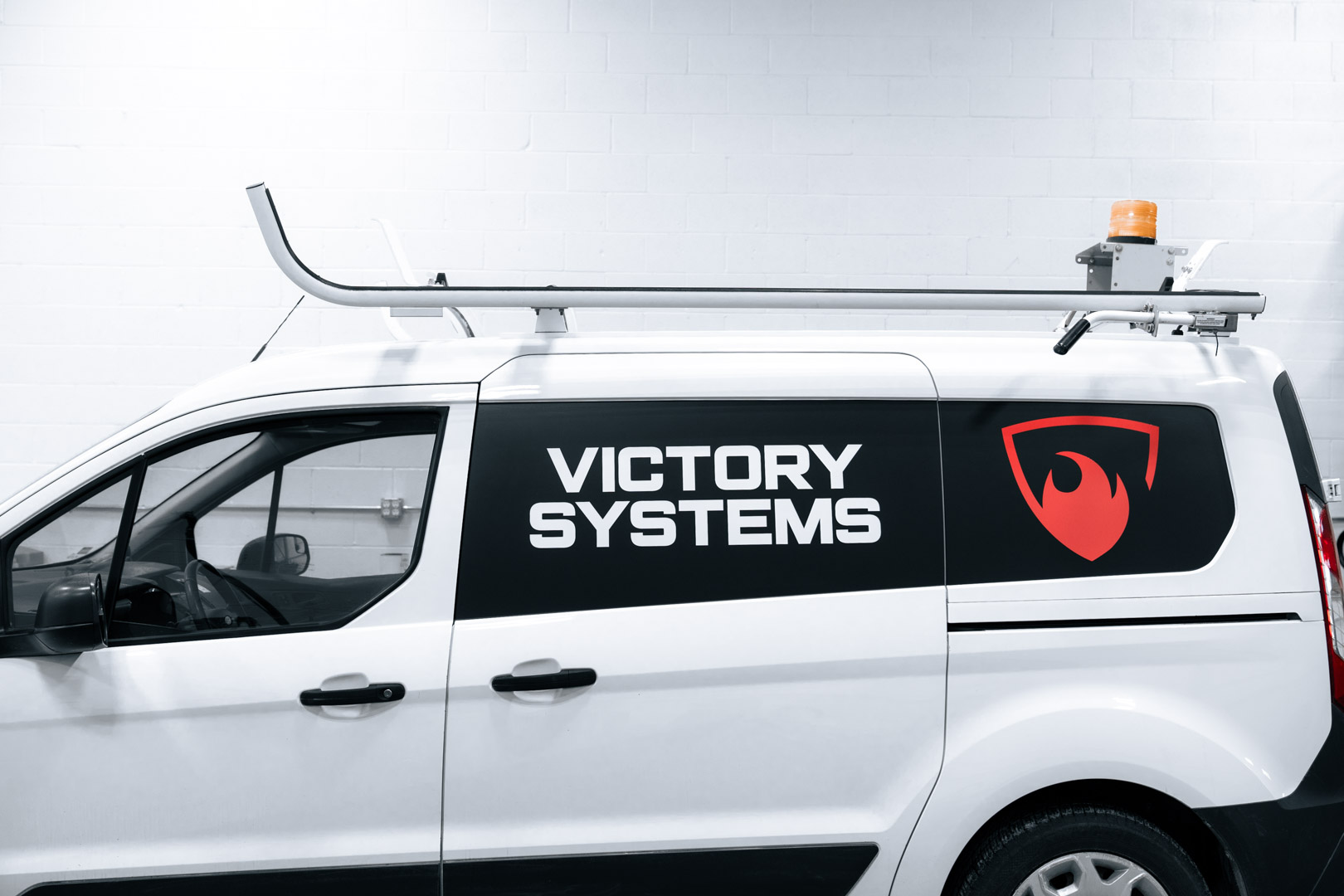 White service van with 'Victory Systems' text and red flame shield logo on the side, equipped with a roof rack and an orange warning light.