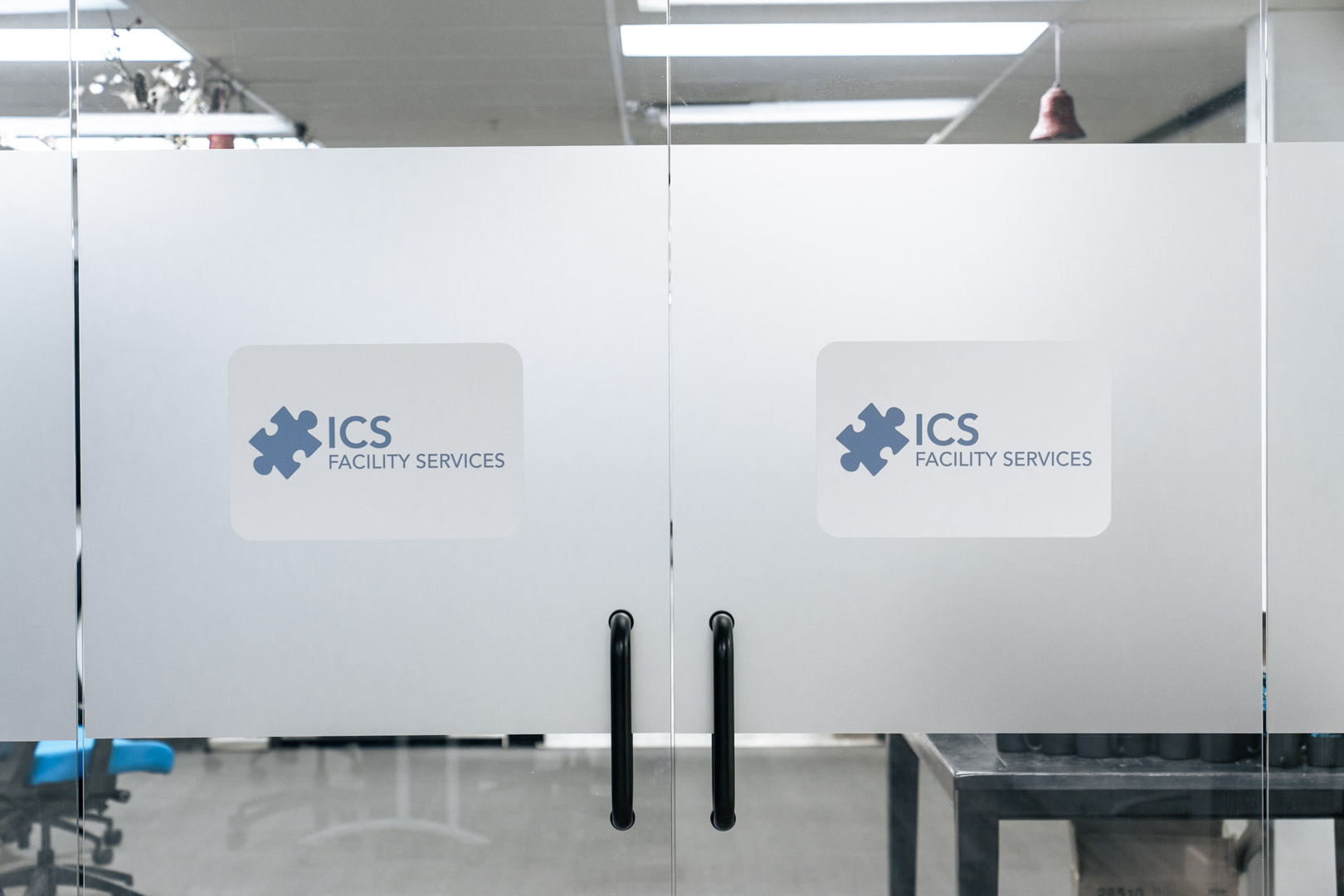 Glass double doors with black handles displaying ICS Facility Services logos on frosted panels.