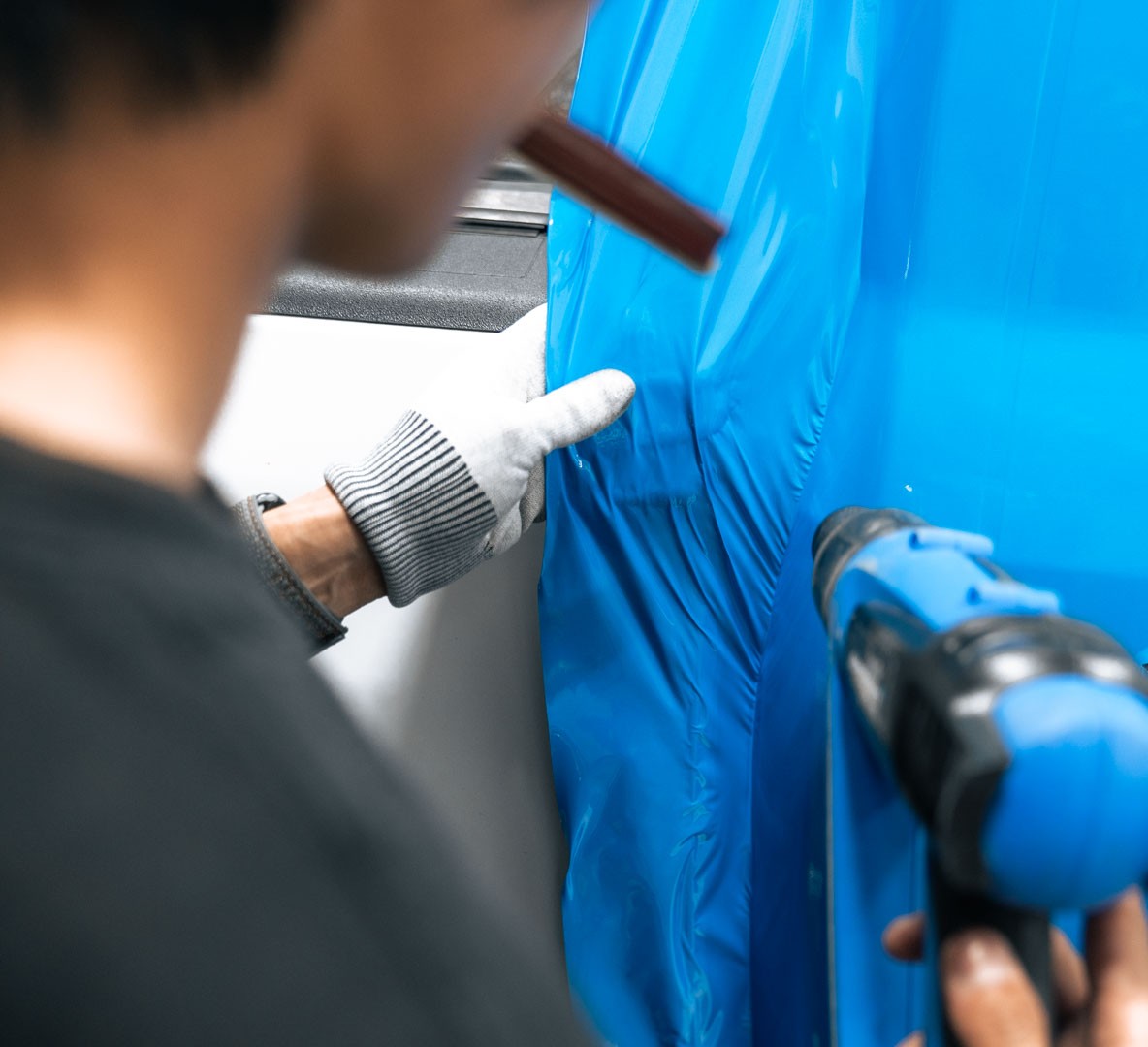 Person wearing white glove using a heat gun to apply or remove blue vinyl wrap on a surface.