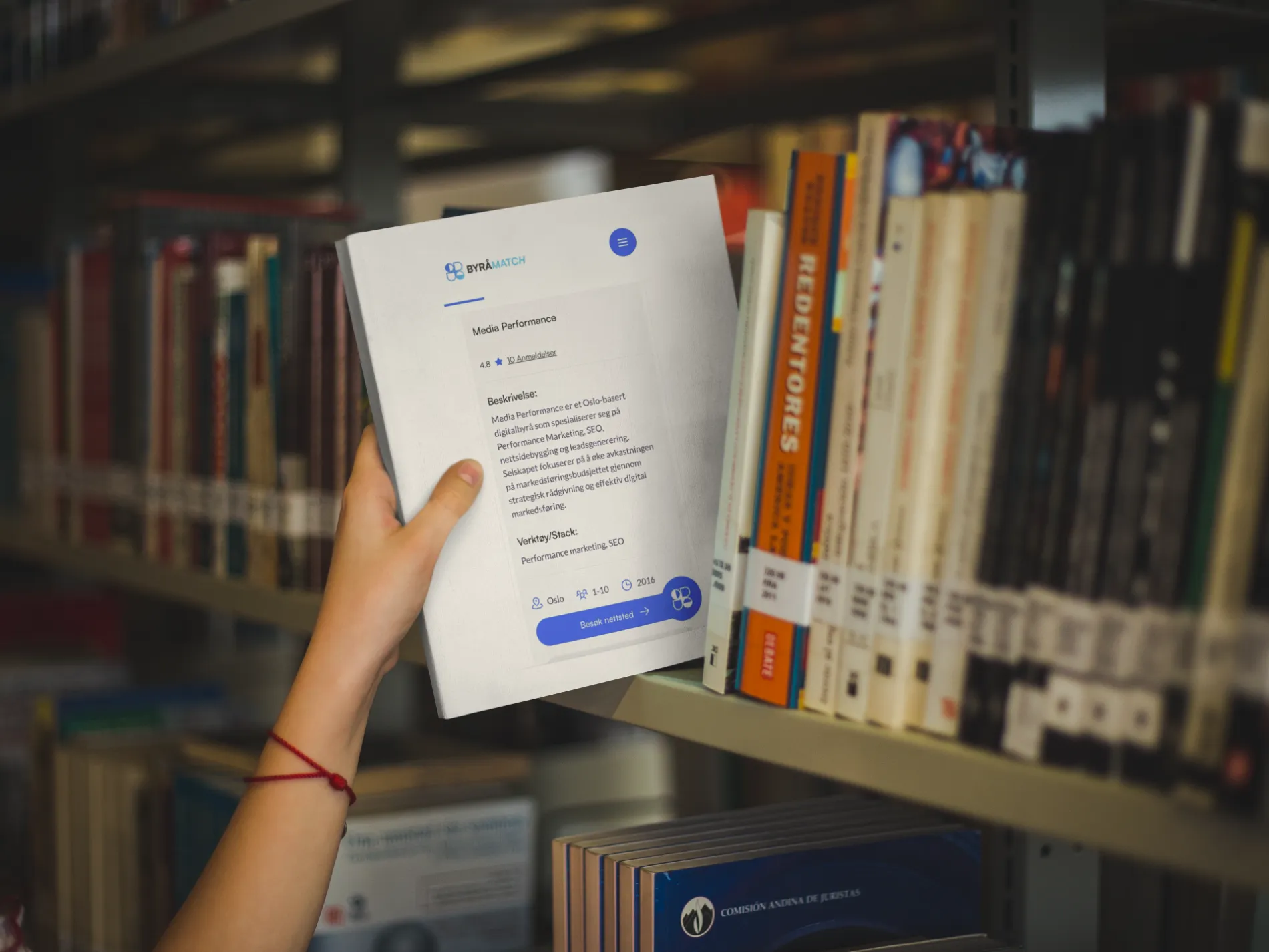 Hånd som holder et papirdokument foran en bokhylle fylt med bøker i et bibliotek.