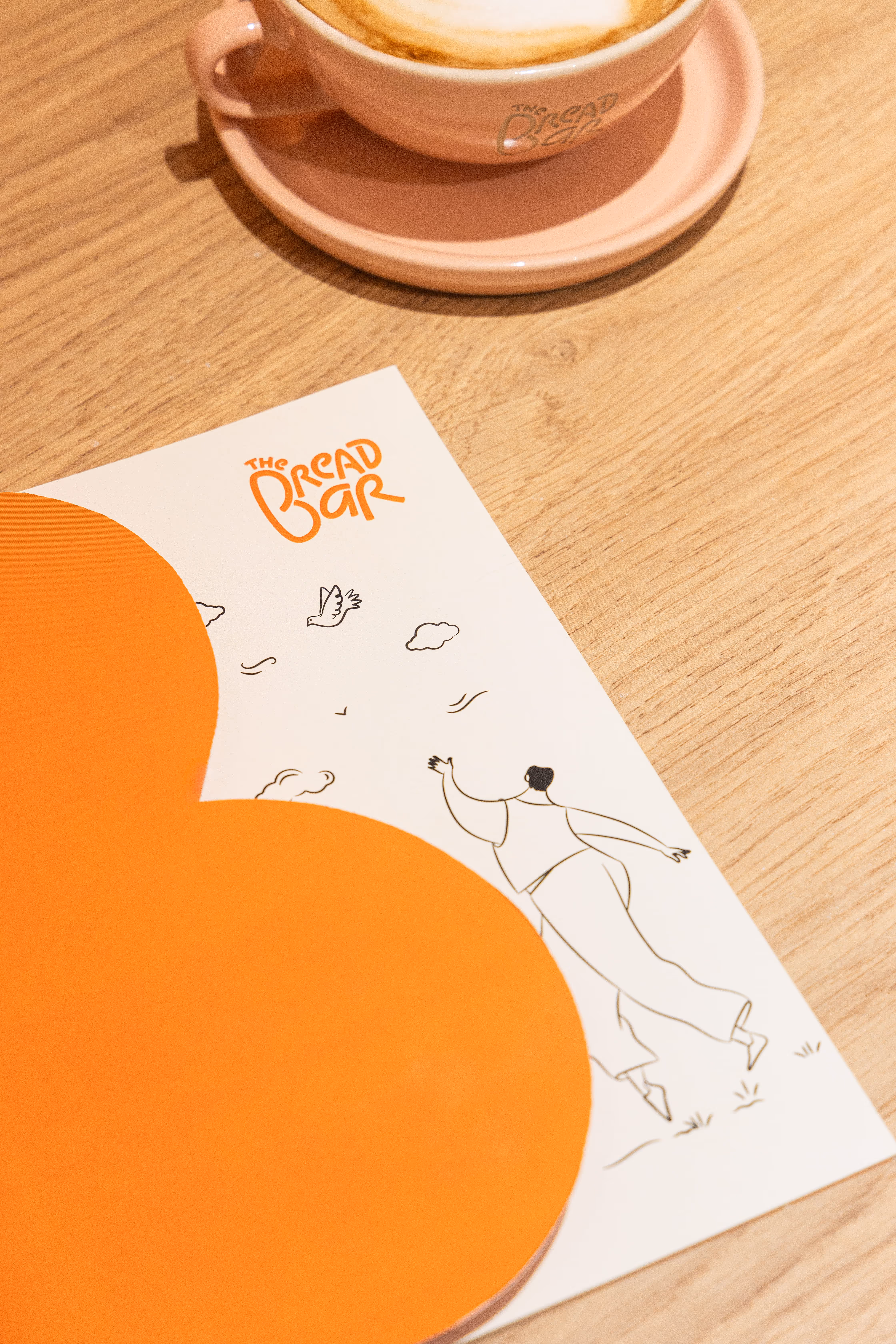 A pink cup of cappuccino on a saucer with a menu from The Bread Bar featuring an orange abstract shape and a line drawing of a person with birds and clouds.