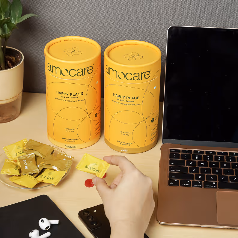 Hand holding a yellow amocare gummy wrapper near two yellow amocare Happy Place supplement containers on a desk with a laptop, phone, earbuds, and a plant.