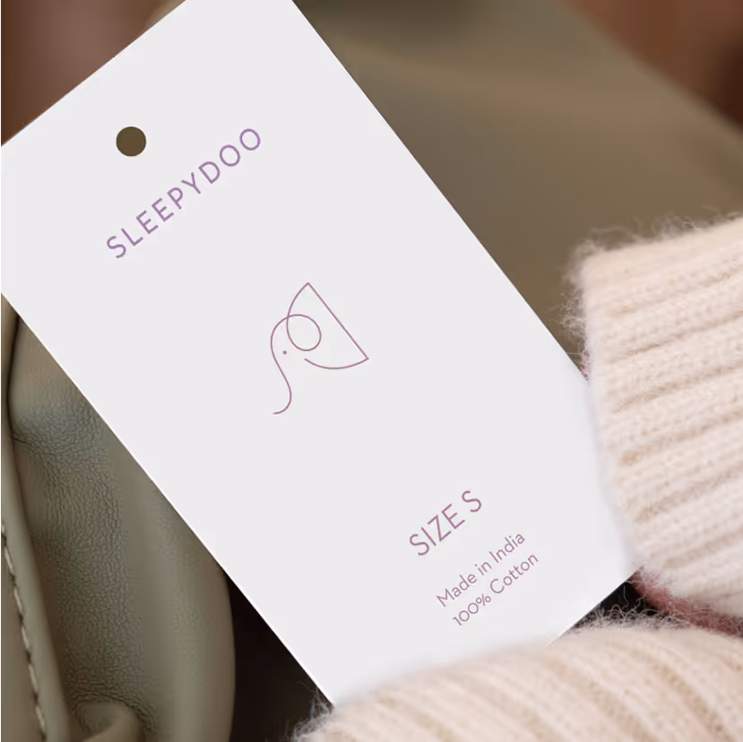 White clothing tag labeled SLEEPYDOO showing size S, made in India, and 100% cotton, held next to a light beige knitted fabric.