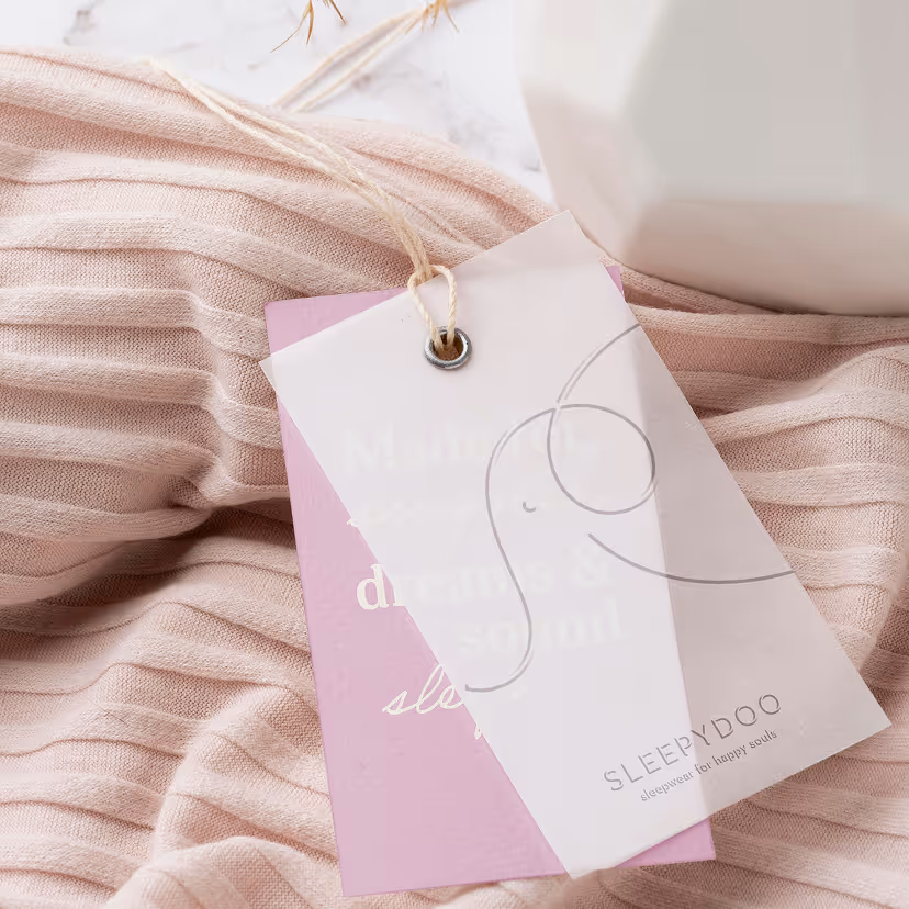 Two clothing tags attached with string, one white with an abstract line drawing and the brand name SLEEPYDOO, resting on a folded pink ribbed fabric.