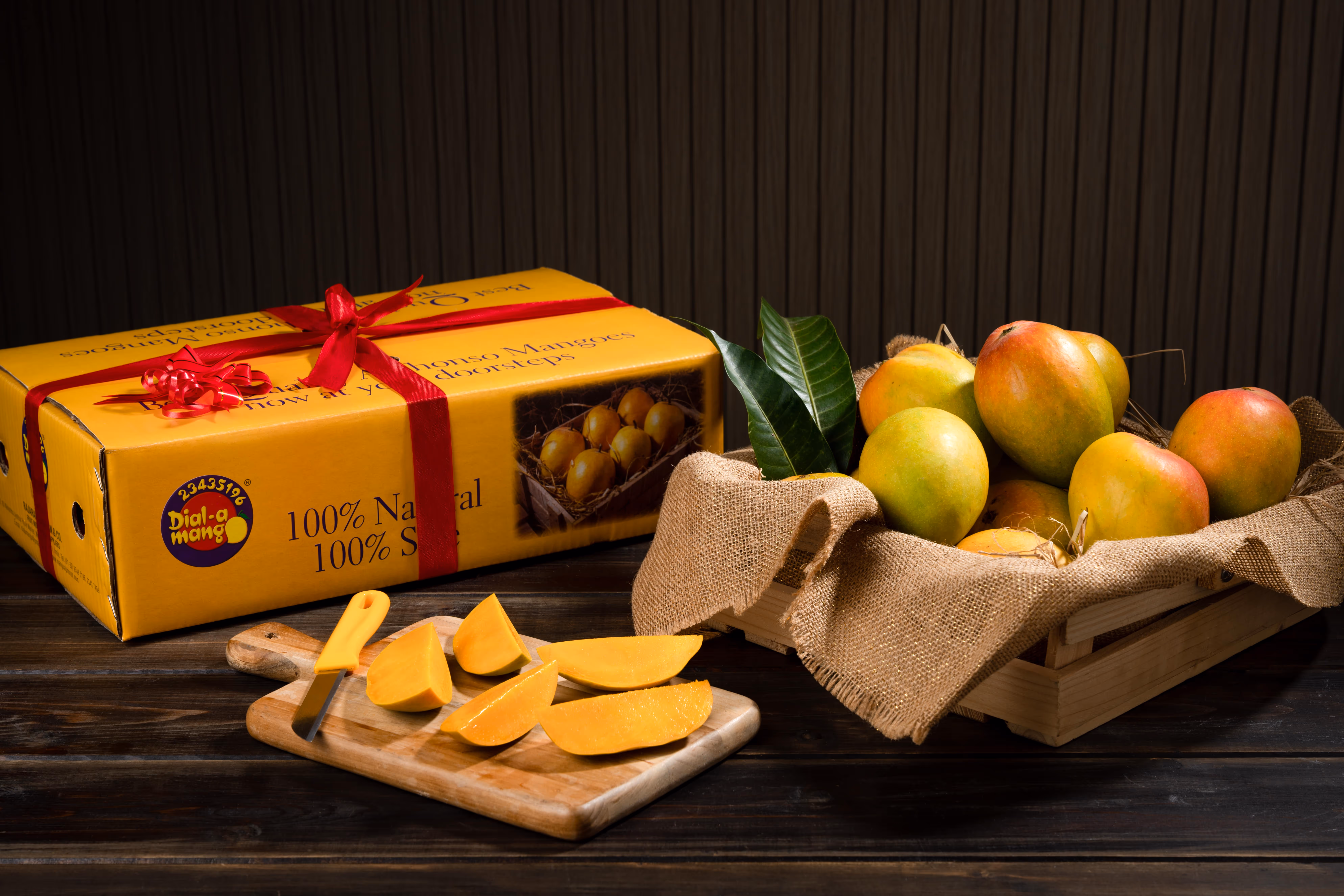 Wooden crate lined with burlap holding ripe mangoes next to a wooden cutting board with sliced mango pieces and a yellow-handled knife, beside a yellow gift box wrapped with a red ribbon.