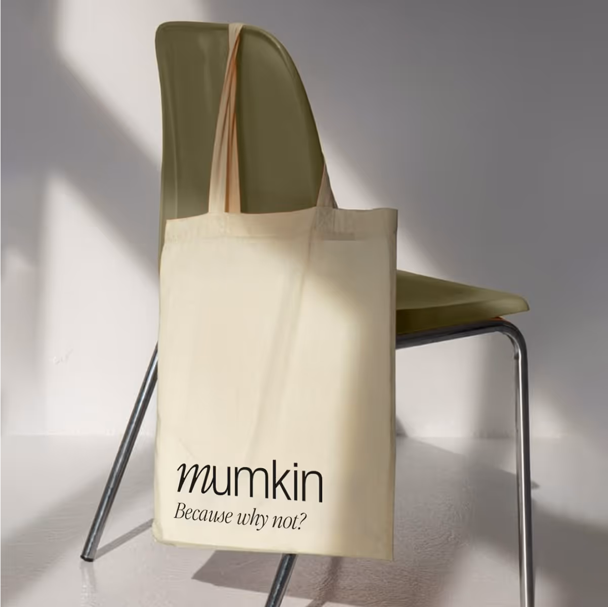 Cream-colored tote bag hanging on the back of an olive green chair with metal legs, printed with the text 'mumkin Because why not?'