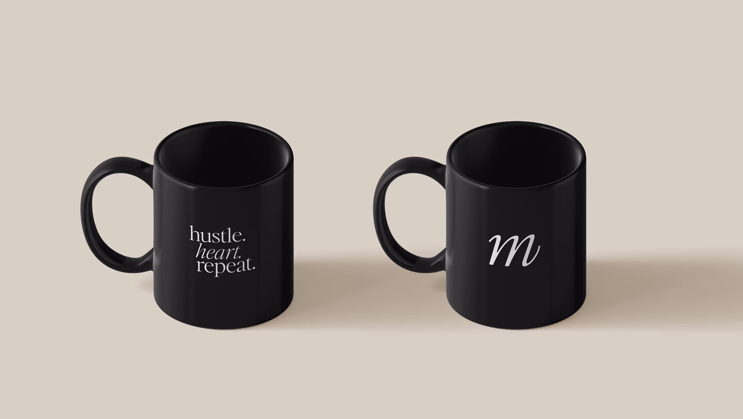 Two black mugs on a beige surface; the left mug reads 'hustle. heart. repeat.' in white text, the right mug displays a white letter 'm'.