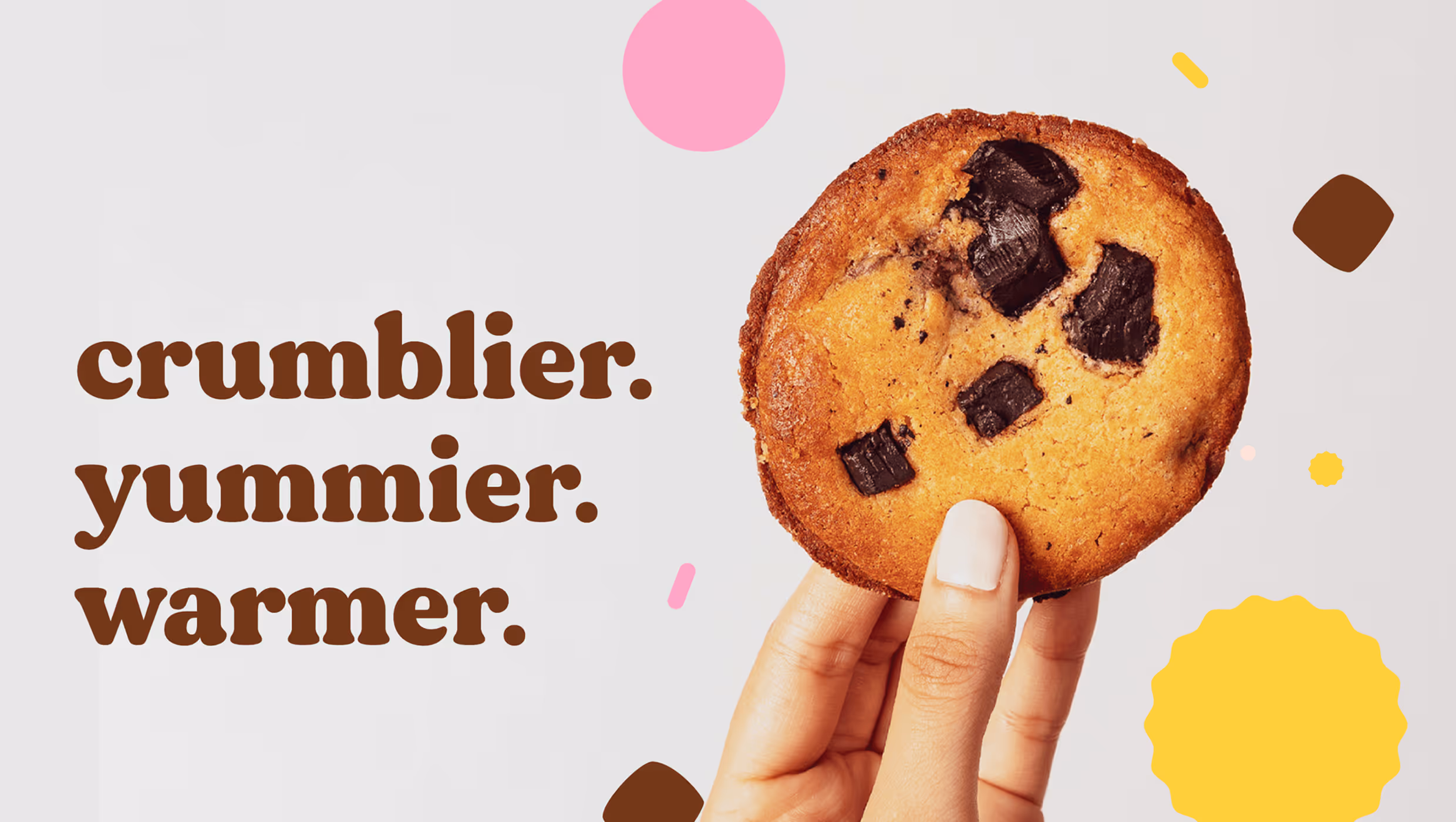 Hand holding a chocolate chip cookie with the text 'crumblier. yummier. warmer.' on a white background with colorful confetti dots.