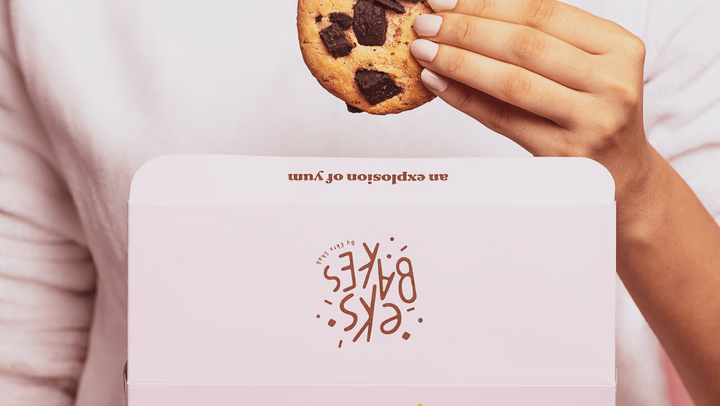 Hand holding a chocolate chip cookie above a pink box labeled 'eks:bakes'.