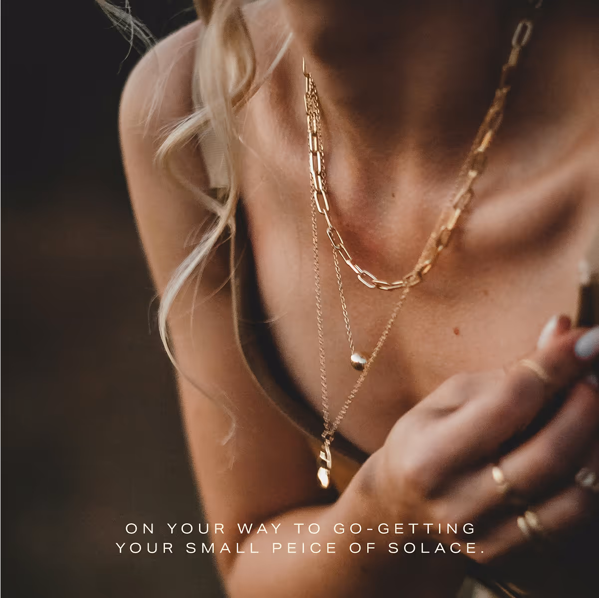 Close-up of a woman wearing layered gold necklaces and rings, with blonde hair curls visible.