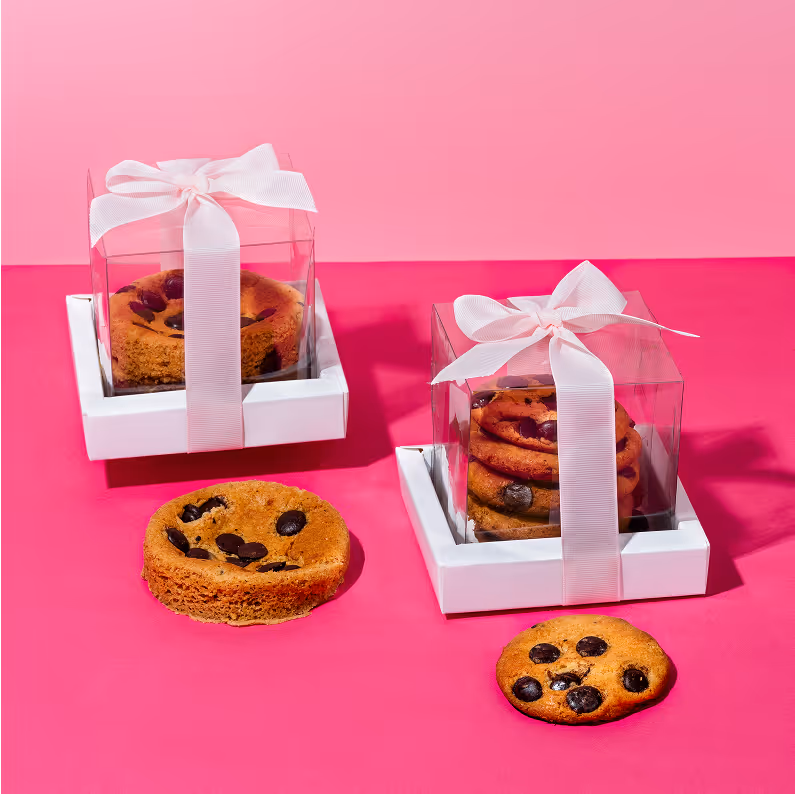 Two stacks of chocolate chip cookies in clear gift boxes with white bases and white ribbons, placed on a pink surface with one cookie outside each box.