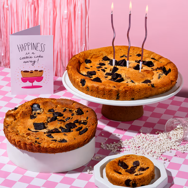Three chocolate chip cookie cakes of different sizes on a pink checkered tablecloth, with one cake on a white cake stand with three lit candles and a card saying 'Happiness is a cookie cake away!'