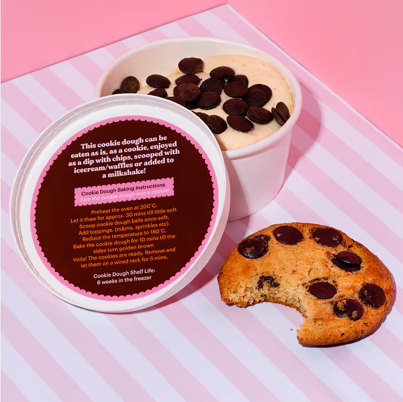 White cup filled with chocolate chip cookie dough topped with chocolate chips alongside a partially eaten baked chocolate chip cookie on a pink and white striped surface.