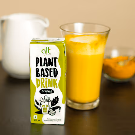 Carton of alt plant-based drink original flavor placed next to a glass of orange-yellow beverage on a dark surface.