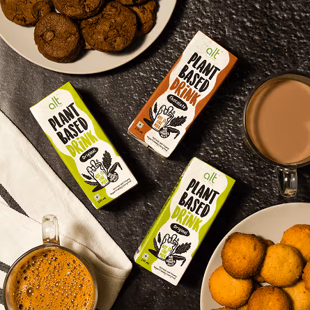 Two green and one brown cartons of alt plant-based drink on a table with plates of cookies and cups of coffee and hot chocolate.