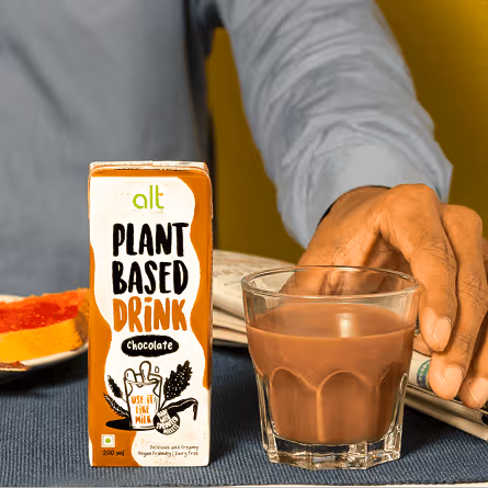A hand reaching for a glass of chocolate plant-based drink next to a carton labeled 'alt Plant Based Drink Chocolate' on a blue placemat.