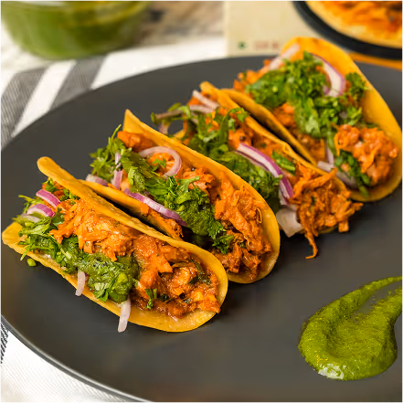 Three tacos filled with shredded meat, chopped cilantro, and sliced red onions on a black plate with green sauce on the side.