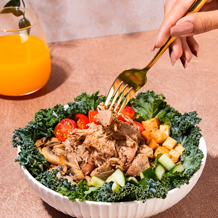 Hand holding a fork lifting tuna from a kale salad bowl with cherry tomatoes, cucumbers, mushrooms, and croutons, next to a glass of orange juice.