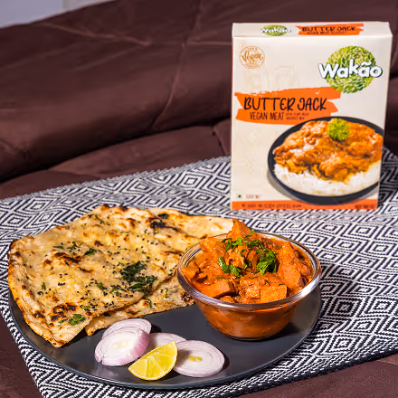 Plate with Indian-style butter jack vegan curry, two pieces of naan bread, sliced red onions, and a lemon wedge, with a Wakao butter jack vegan meat sauce box in the background.