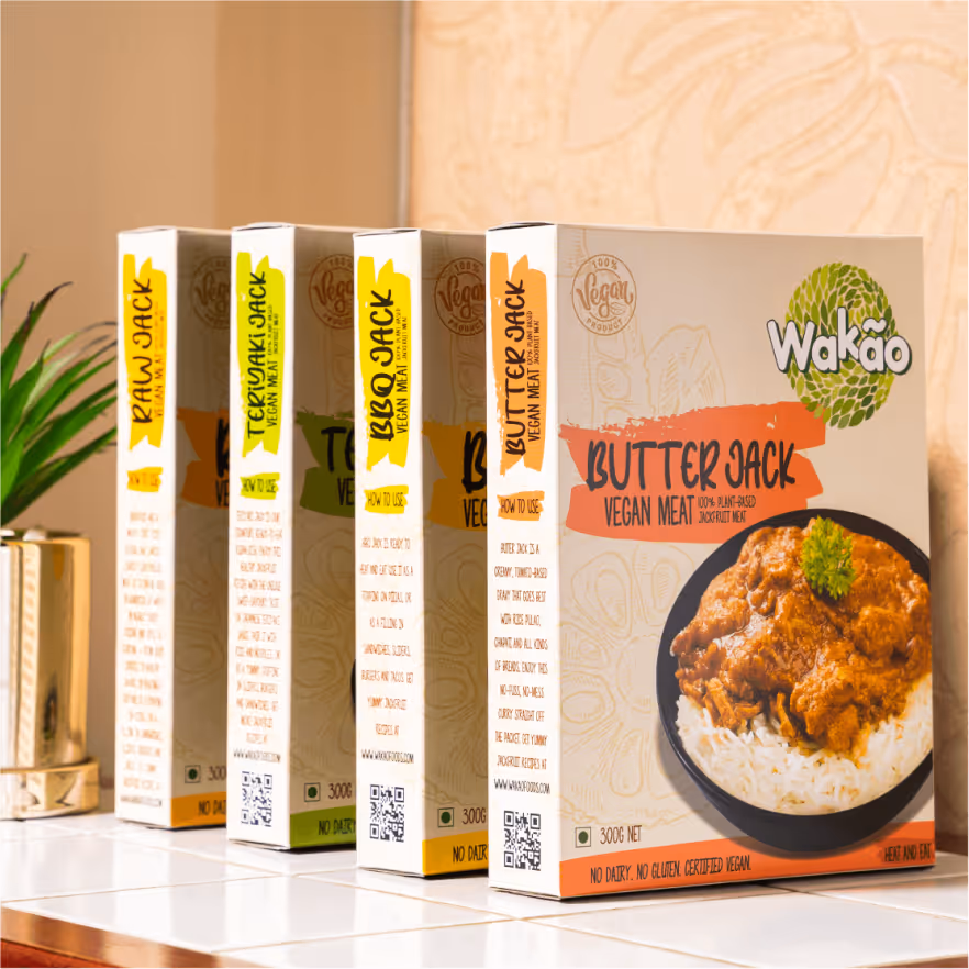 Four boxes of Wakaõ vegan meat products including Butter Jack, Raw Jack, and Teriyaki Jack displayed on a white tiled surface with a green plant in a gold pot in the background.