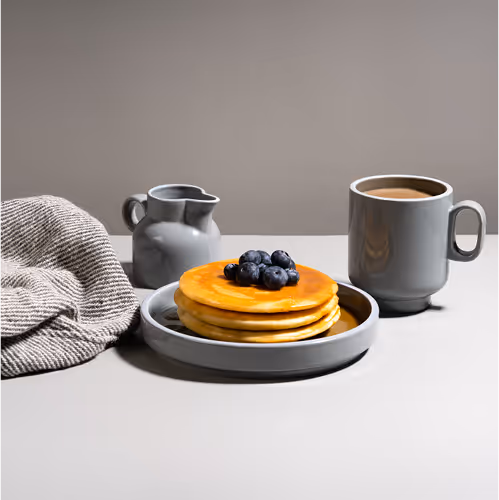 Stack of three pancakes topped with blueberries on a gray plate next to a gray cup of coffee and a small gray pitcher on a light gray table with a folded beige cloth.
