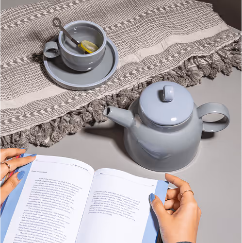 Hands with blue painted nails holding an open book next to a gray teapot and a gray cup with a tea bag on a matching saucer, placed on a gray table with a textured cloth.