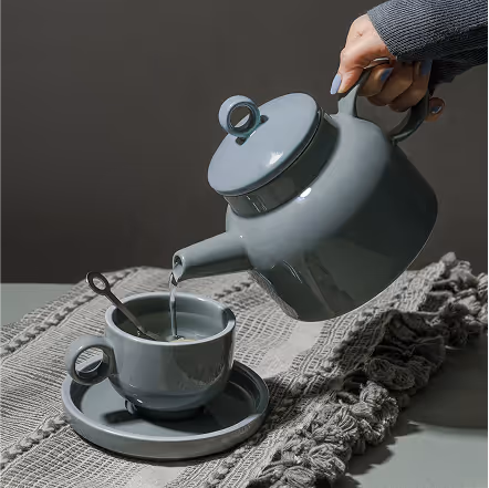 Hand pouring tea from a gray teapot into a matching cup on a gray textured cloth.