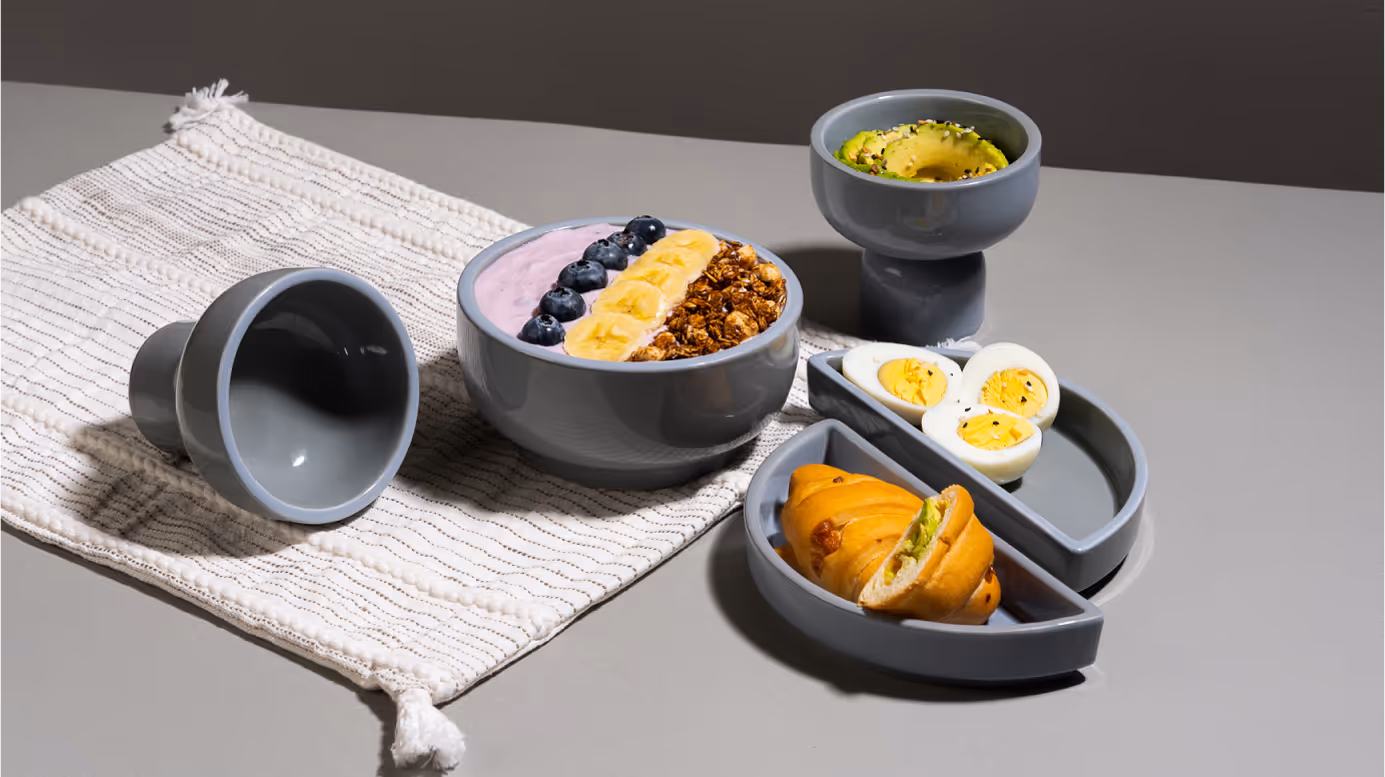 Breakfast spread with a bowl of smoothie topped with blueberries, banana, and granola, sliced avocado in a pedestal bowl, halved boiled eggs, and a croissant on segmented gray dishes with a striped cloth.