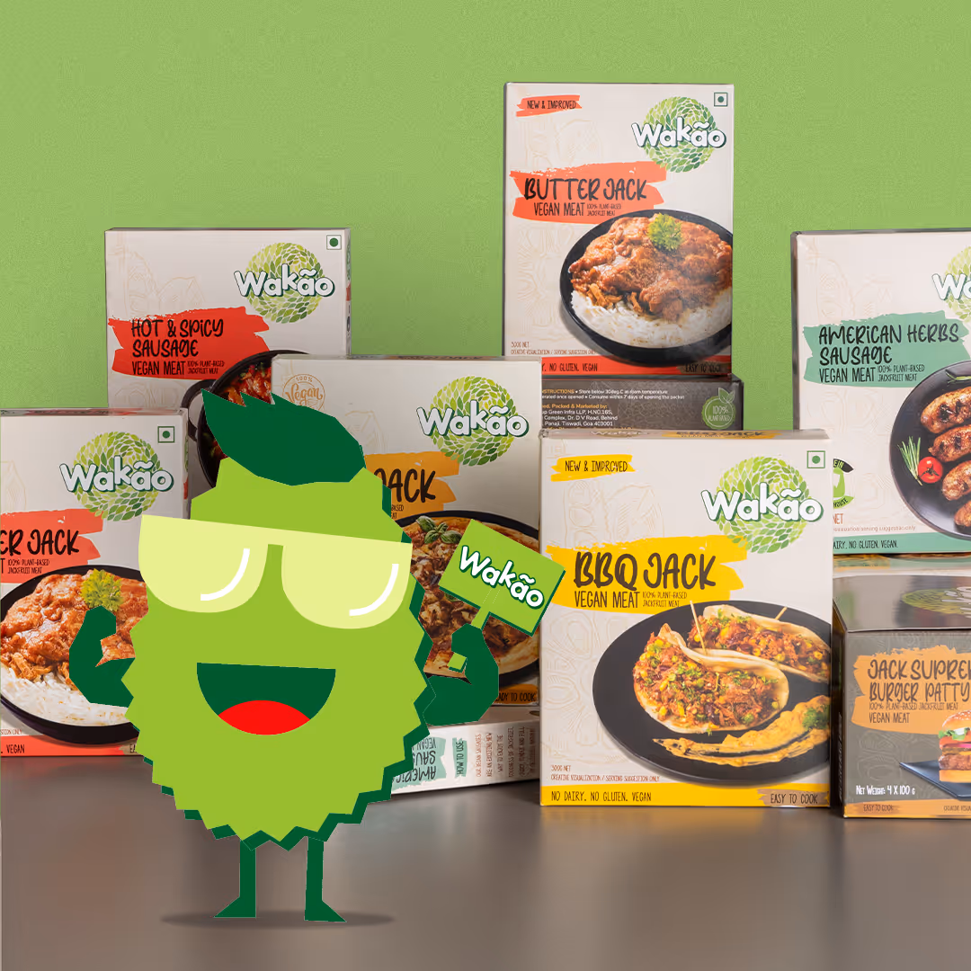 Boxes of Wakao branded vegan meat products including Butter Jack, Hot & Spicy Sausage, BBQ Jack, and American Herbs Sausage arranged on a table with a cartoon green character holding a Wakao sign in front.