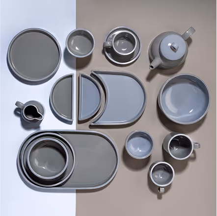 Top-down view of an assortment of modern gray ceramic tableware including plates, bowls, mugs, and a teapot arranged on a split white and beige background.