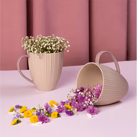 Two beige mugs with ribbed texture; one upright filled with small white flowers, the other tipped over spilling purple and yellow flowers on a light pink surface.