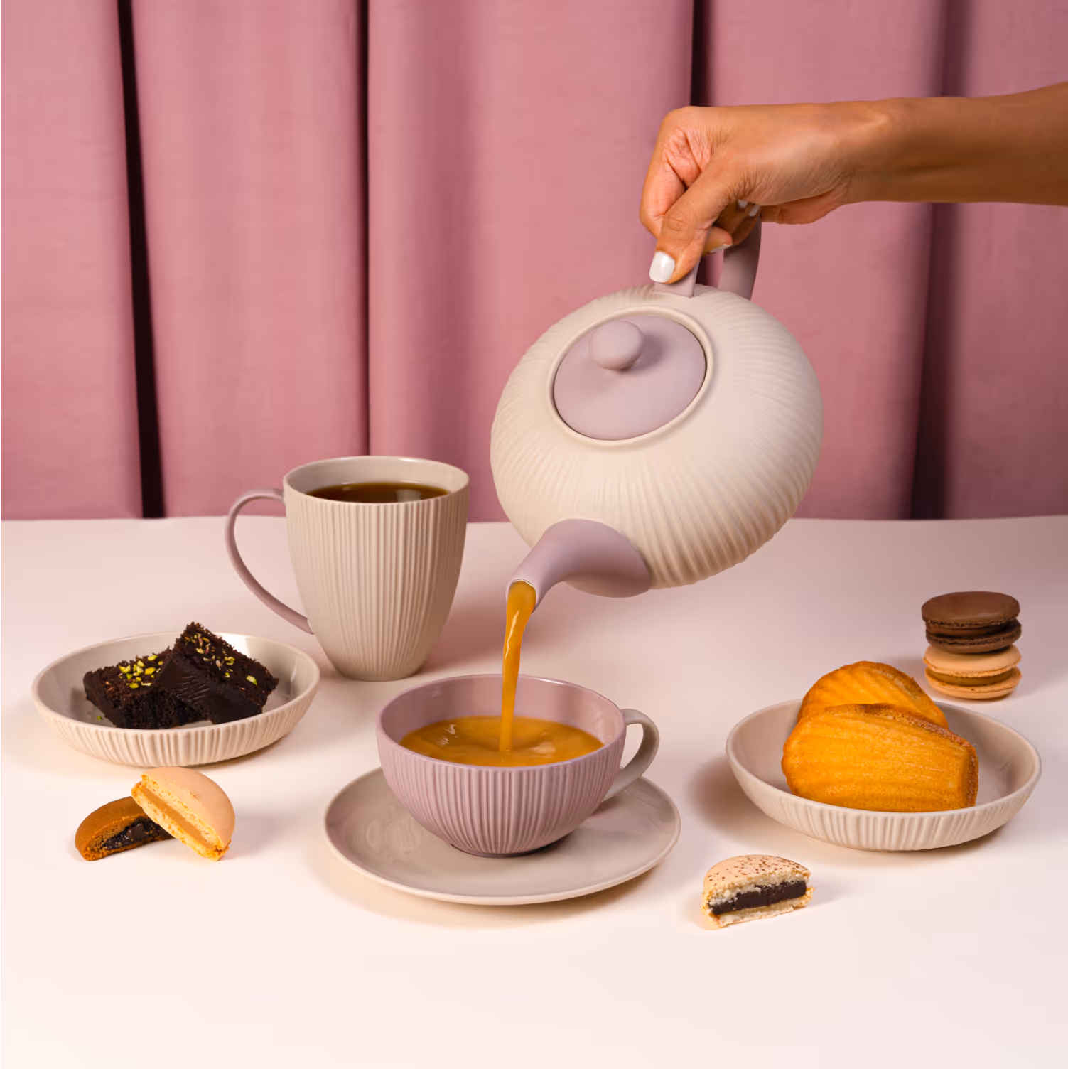 Hand pouring tea from a ribbed teapot into a pink teacup surrounded by desserts on a white table with a pink curtain background.