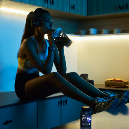 Woman in workout attire wearing headphones and drinking from a bottle while sitting on kitchen counter with fitness supplement container in foreground.
