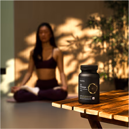 Black bottle of XS Omega supplement on a wooden table with a woman meditating in the background.