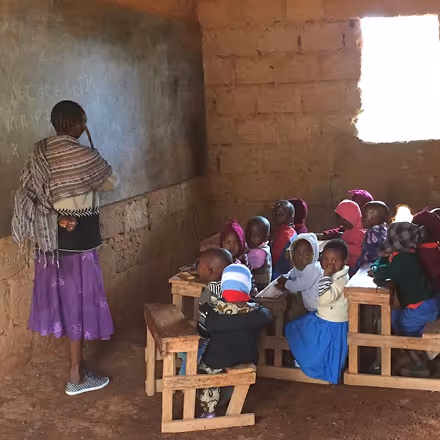 Teacher standing by a chalkboard in a simple classroom with young students sitting at wooden desks.